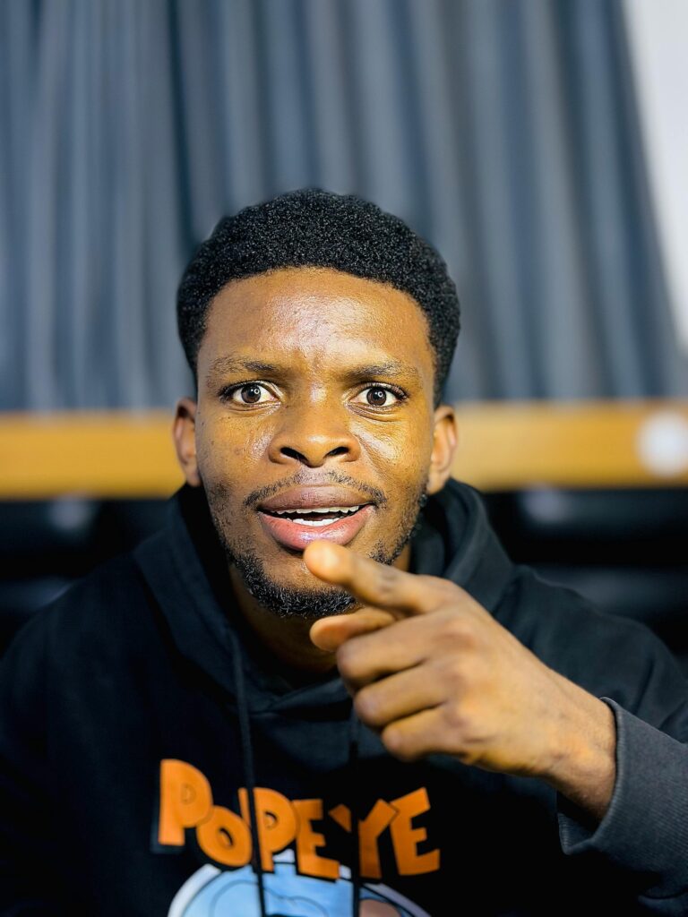 A young man in a hoodie looks surprised while pointing at the camera indoors.